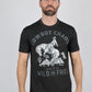 A man wearing a black graphic t-shirt with a white western print of a cowboy on a bucking horse and the text 'COWBOY CHAOS love to be WILD AND FREE', paired with blue jeans, against a solid gray background.