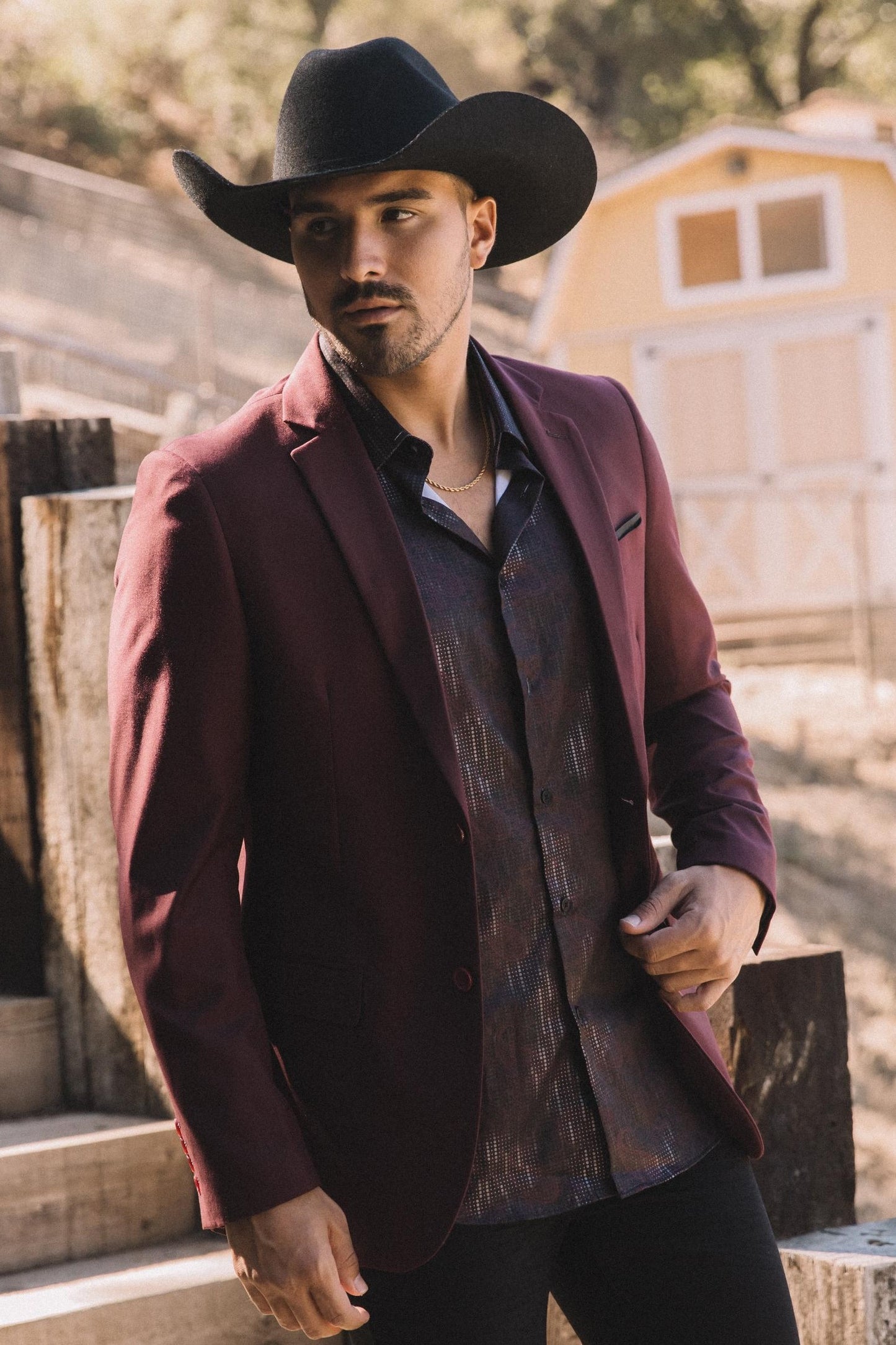 Man wearing a burgundy blazer and cowboy hat in a rustic setting