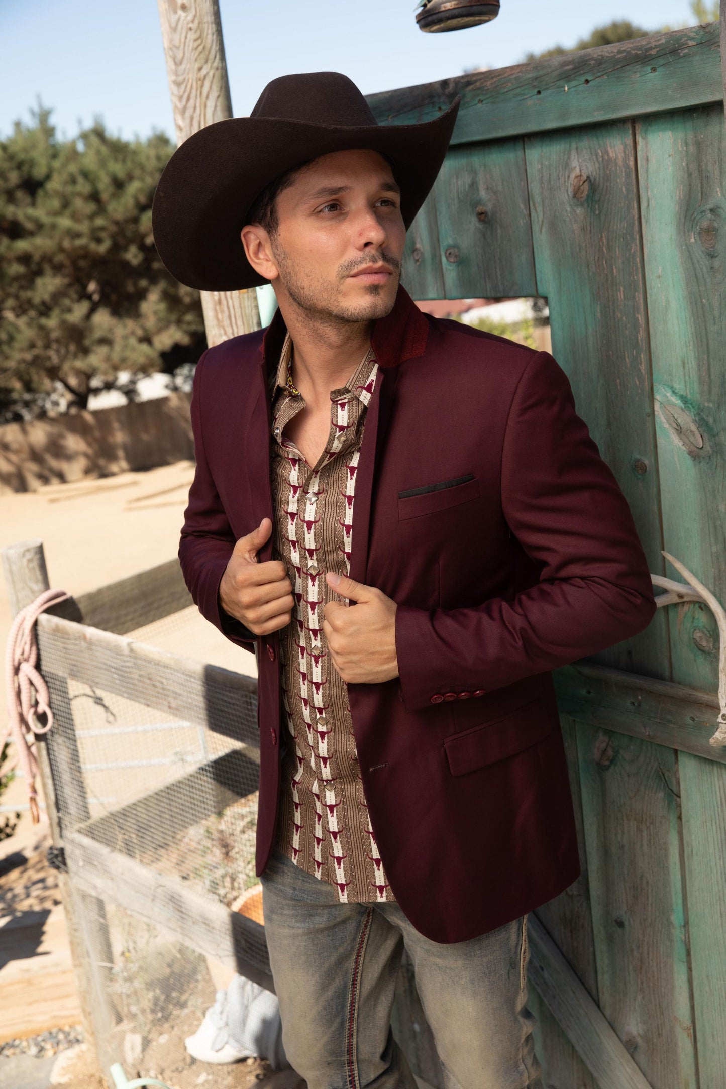 Man wearing a burgundy blazer and cowboy hat standing next to a wooden fence.