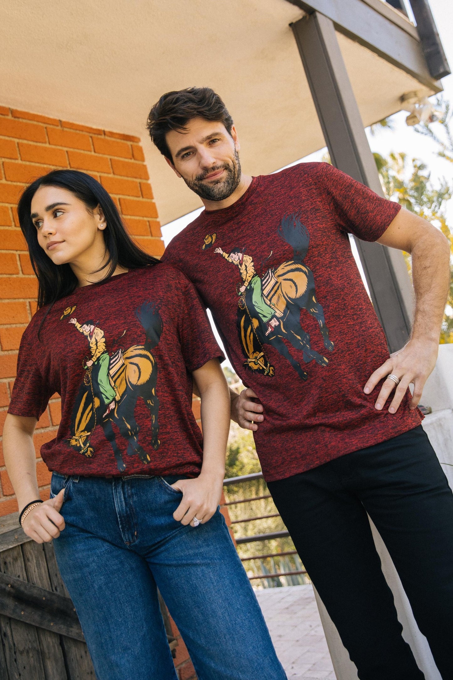 A man and a woman modeling matching heathered burgundy t-shirts with a graphic print of a cowboy riding a horse, posed outdoors on a porch.