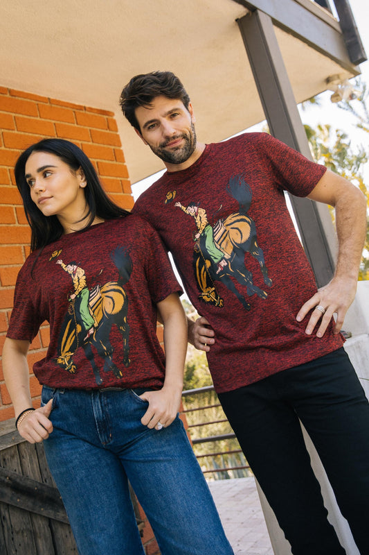 A man and a woman modeling matching heathered burgundy t-shirts with a graphic print of a cowboy riding a horse, posed outdoors on a porch.